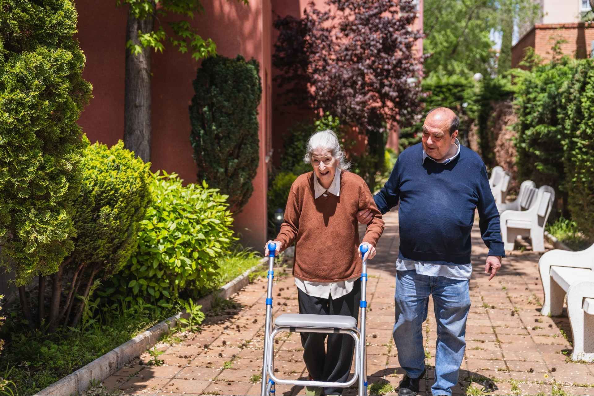 A senior woman with gray hair using a walker, assisted by a senior man walking beside her on a paved path lined with greenery in a residential care setting.
